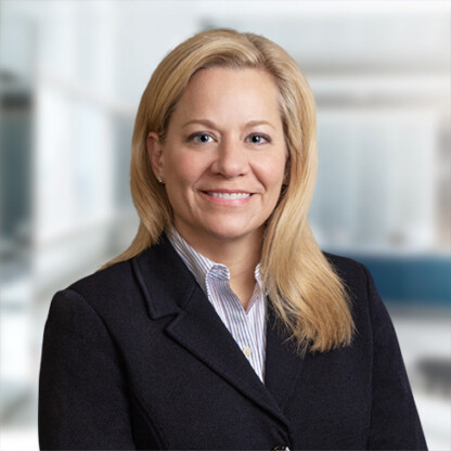 A woman with straight blonde hair wearing a dark blazer and striped shirt smiles at the camera in a bright corporate law office setting.