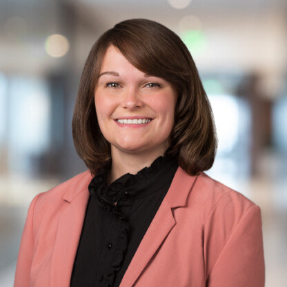 A woman with brown hair, wearing a pink blazer and black blouse, smiles while posing in a softly blurred indoor corporate law office setting.