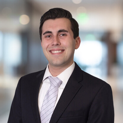 A young man in a dark suit and light tie smiles at the camera, standing in a brightly lit, blurred office—a professional setting often seen in law offices with Chicago lawyers.