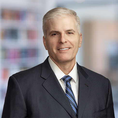 A man in a dark suit and striped tie stands in front of a blurred background with shelves and books, reflecting the professional atmosphere of a corporate law office.