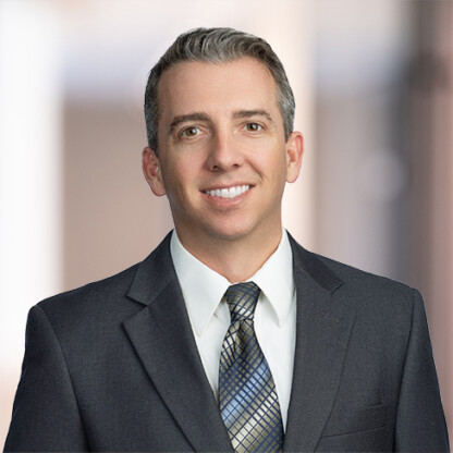 A man in a dark suit, white shirt, and patterned tie smiles at the camera against a blurred indoor background, reflecting the professionalism often found in law offices specializing in intellectual property law.