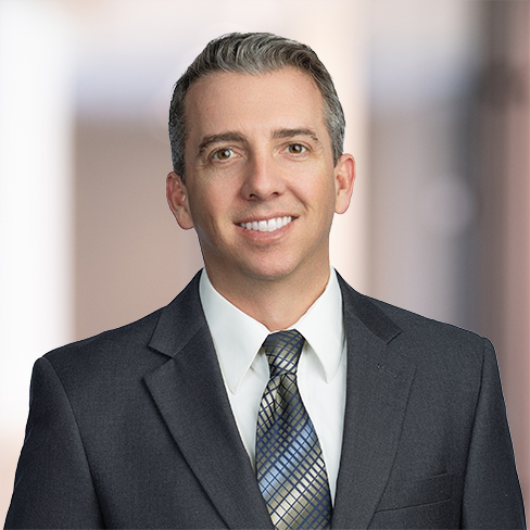 A man in a dark suit, white shirt, and patterned tie smiles at the camera against a blurred indoor background, reflecting the professionalism often found in law offices specializing in intellectual property law.