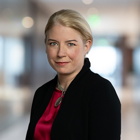 A woman with blonde hair pulled back, wearing a black blazer and a red blouse, stands in a blurred corporate law office hallway.
