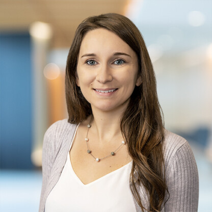 A woman with long brown hair, wearing a light purple cardigan, white top, and beaded necklace, smiles in a brightly lit, blurred office setting at one of the leading law offices providing litigation support.
