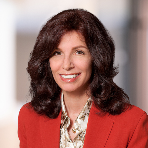 A woman with medium-length wavy brown hair, wearing a red blazer over a patterned blouse, smiles at the camera in a softly blurred indoor setting, representing one of the top law offices for lawyers in Chicago.