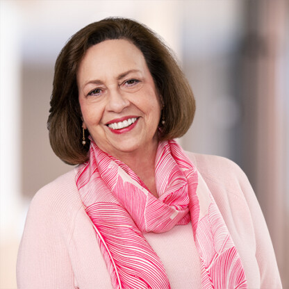 A middle-aged woman with shoulder-length brown hair, wearing a light pink top and a pink patterned scarf, smiles at the camera against a softly blurred indoor background—perfect for profiles of lawyers in Chicago or intellectual property law experts.