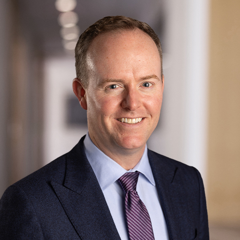 A man in a suit and tie stands indoors, smiling at the camera with a blurred hallway background in a leading corporate law office.