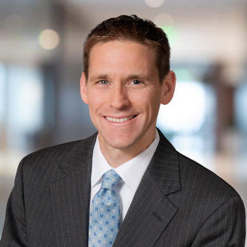 A man in a suit and tie smiles at the camera with a blurred corporate law office in the background.