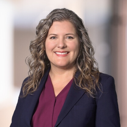 Woman with wavy, shoulder-length gray hair wearing a dark blazer over a burgundy top, smiling at the camera—a confident presence among leading lawyers in Chicago, specializing in intellectual property law. Blurred indoor background.