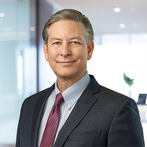 A middle-aged man with gray hair in a suit and red tie stands in a modern corporate law office, smiling at the camera.