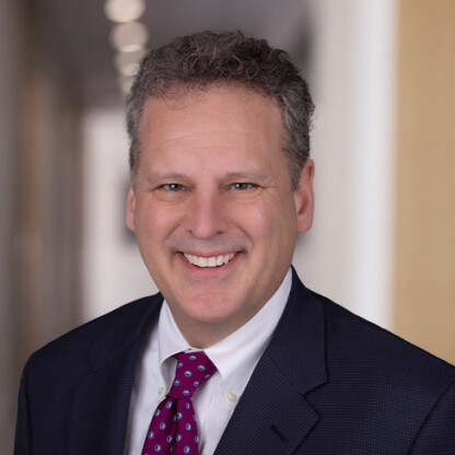 A middle-aged man with curly gray hair, wearing a dark suit, white shirt, and red patterned tie, smiles at the camera in a brightly lit corporate law office.