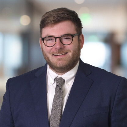A man wearing glasses, a blue suit, white shirt, and patterned tie stands indoors with a blurred corporate law office background.