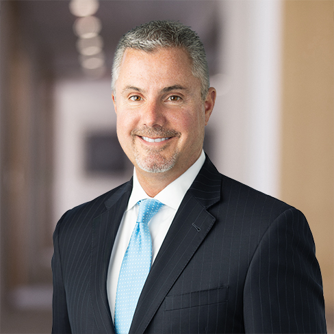 A man in a dark pinstripe suit and light blue tie stands smiling in a blurred law offices hallway background.