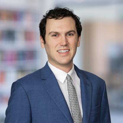 A man in a blue suit and patterned tie smiles at the camera, standing in a corporate law office with blurred bookshelves in the background.