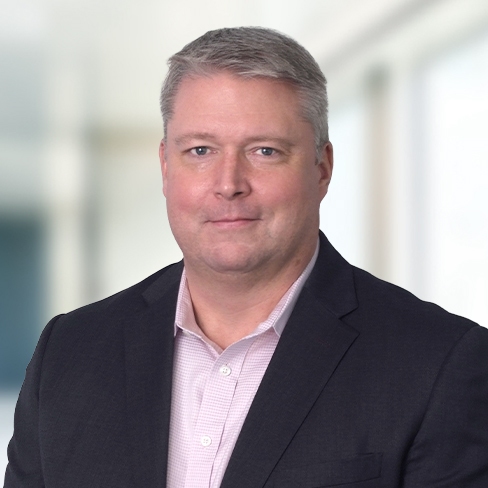 A man with short gray hair wearing a dark suit jacket and light pink collared shirt stands in front of a blurred indoor background, reflecting the professional atmosphere of a corporate law office.