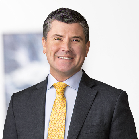 A man in a dark suit and yellow patterned tie smiles in front of a blurred white and blue background, reflecting the professional atmosphere of a corporate law office.