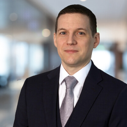 A man in a dark suit, white shirt, and patterned tie stands in a corporate law office with a blurred background, exuding professionalism.