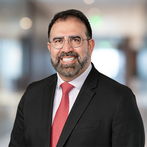 A man with glasses and a beard, wearing a black suit, white shirt, and red striped tie stands in a blurred corporate law office setting.
