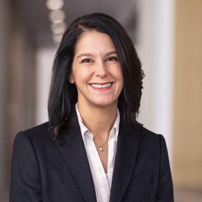 A woman with dark shoulder-length hair wearing a navy blazer and white blouse stands indoors, smiling at the camera with a blurred hallway background—an inviting scene often found in top law offices or among leading lawyers in Chicago.