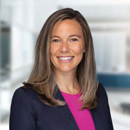 A woman with straight, shoulder-length light brown hair smiling, wearing a dark blazer over a bright pink top, in a modern corporate law office setting.