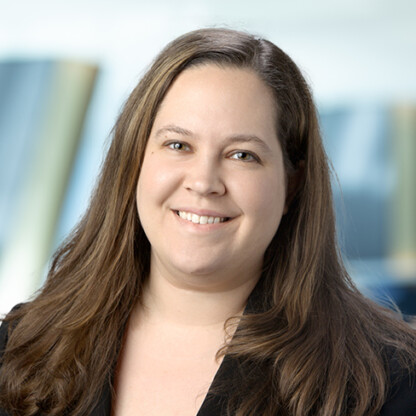 A woman with long brown hair and a black blazer smiles at the camera against a blurred blue and beige background, embodying the confidence of top Chicago lawyers.
