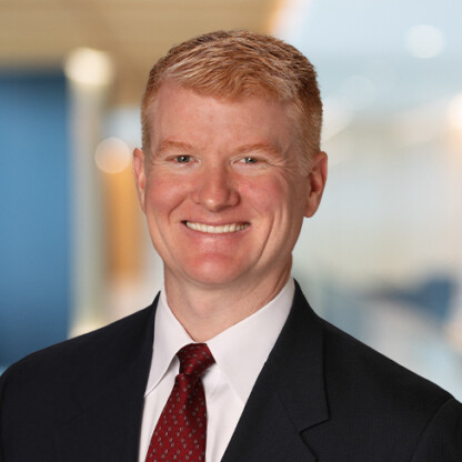 A man with short red hair wearing a dark suit, white shirt, and red tie, smiling in an office setting with a blurred background, representing lawyers in Chicago specializing in intellectual property law.