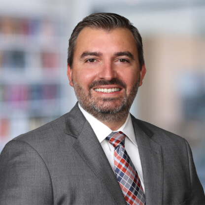 A man in a gray suit, white shirt, and plaid tie smiles at the camera in a corporate law office, with a blurred background—ideal for those seeking skilled lawyers in Chicago.