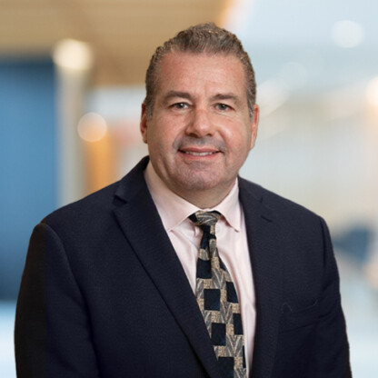 A middle-aged man in a suit and patterned tie poses for a professional headshot against a blurred office background, reflecting the polished image often seen in corporate law office environments.