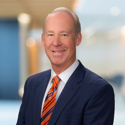 A middle-aged man in a blue suit with an orange striped tie, smiling, posed in a corporate law office setting with a blurred background.