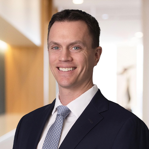 A man in a dark suit and light blue tie is smiling, standing in a well-lit corporate law office hallway with neutral-colored walls.