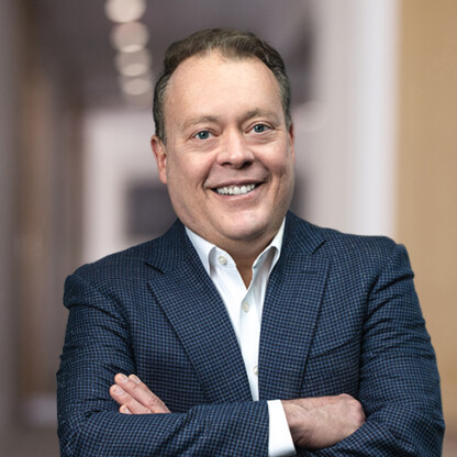 A man in a blue checked suit jacket and white shirt stands with arms crossed, smiling, in a blurred indoor hallway, exuding the professionalism found at top corporate law offices specializing in litigation support.