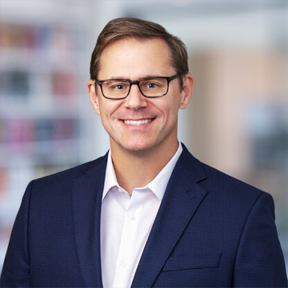 A man wearing glasses, a navy blue blazer, and a white shirt smiles at the camera. The blurred background hints at a corporate law office, perfect for showcasing top lawyers in Chicago.