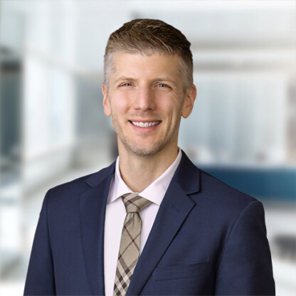 A man in a navy suit and plaid tie smiles in front of a blurred corporate law office background.
