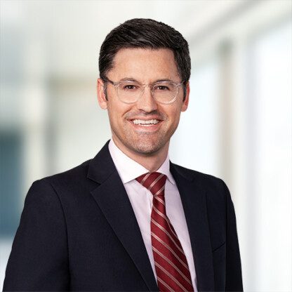 A man in a suit, light pink shirt, and red striped tie, with short dark hair and glasses, stands smiling in a bright, blurred corporate law office setting.