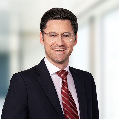 A man in a suit, light pink shirt, and red striped tie, with short dark hair and glasses, stands smiling in a bright, blurred corporate law office setting.