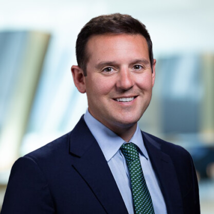 A man in a dark suit, light blue shirt, and green patterned tie smiles at the camera with a blurred corporate law office in the background.