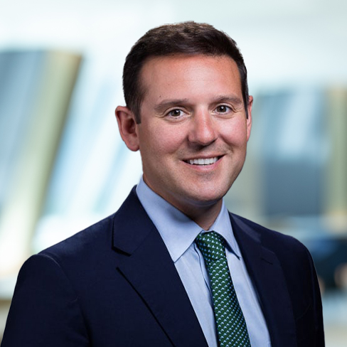 A man in a dark suit, light blue shirt, and green patterned tie smiles at the camera with a blurred corporate law office in the background.