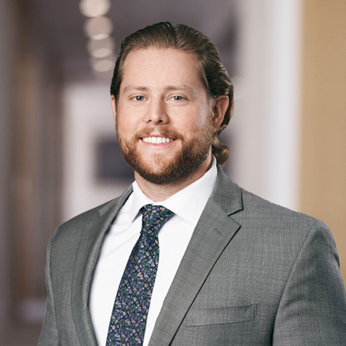 A man with long, light brown hair and a beard, wearing a gray suit, white shirt, and patterned tie, stands in a blurred hallway of a corporate law office.