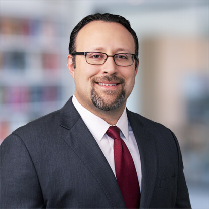 A man wearing glasses, a dark suit, white shirt, and red tie is smiling at the camera in an office setting with blurred bookshelves in the background of renowned Chicago lawyers’ law offices.