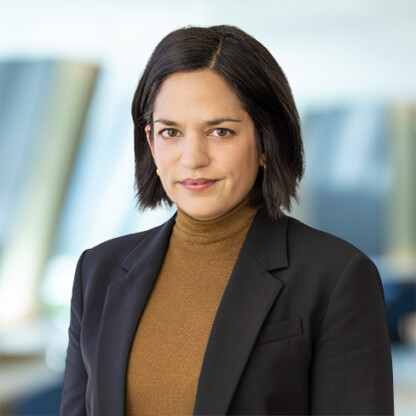 A woman with straight dark hair, wearing a black blazer and brown turtleneck, stands indoors in front of a blurred corporate law office background.