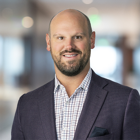 A man with a shaved head and beard, wearing a checked shirt and dark blazer, stands in a blurred office setting—typical of Chicago lawyers specializing in intellectual property law.