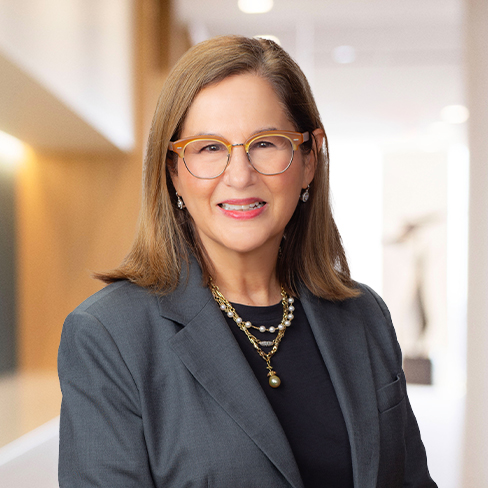 Woman with light brown hair wearing glasses, a dark blazer, black top, and layered necklaces, smiling in a brightly lit corporate law office hallway.
