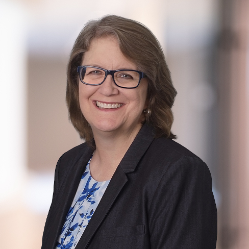 A woman with shoulder-length brown hair and glasses, wearing a dark blazer over a blue and white patterned top, smiles at the camera against a blurred background—representing skilled lawyers in Chicago.