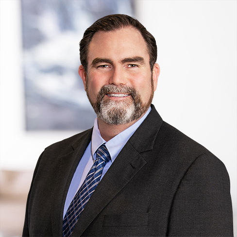 A man with dark hair and a salt-and-pepper beard, wearing a dark suit, light blue shirt, and striped tie, poses for a professional headshot against a blurred background in a corporate law office.