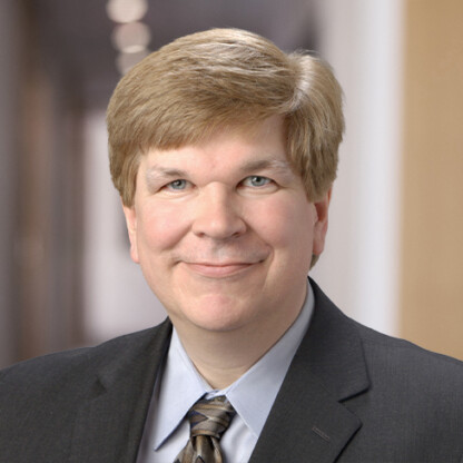 A middle-aged man with light brown hair wearing a dark suit, light blue shirt, and striped tie poses in a blurred office hallway, representing one of the top law offices offering litigation support for lawyers in Chicago.