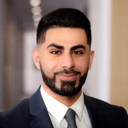 A man with short black hair and a trimmed beard, wearing a dark suit, white shirt, and tie, stands in a hallway with blurred lights—a professional often sought for intellectual property law or litigation support.