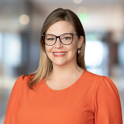 Woman with light brown hair, wearing glasses and an orange blouse, smiles at the camera in a corporate law office with a blurred background.