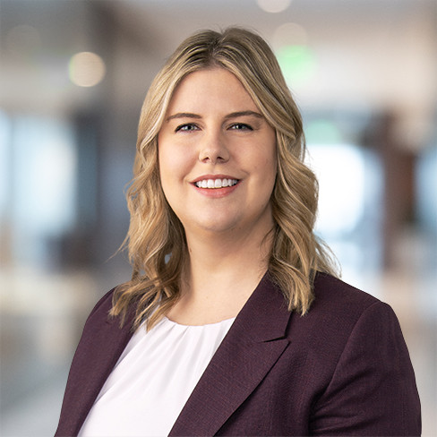 A woman with blonde hair, wearing a white top and maroon blazer, smiles in a brightly lit corporate law office.