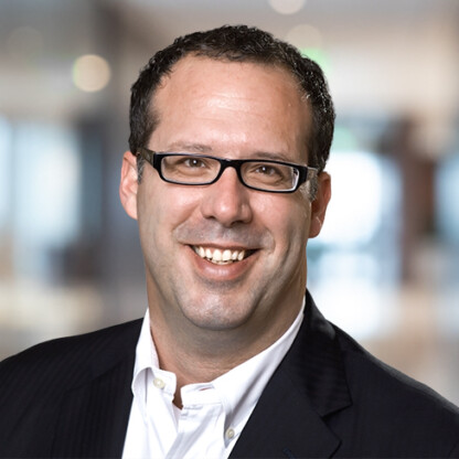 A man with short dark hair and glasses, wearing a dark suit jacket and white shirt, smiles in a blurred indoor setting, reflecting the professionalism often seen in law offices specializing in intellectual property law.