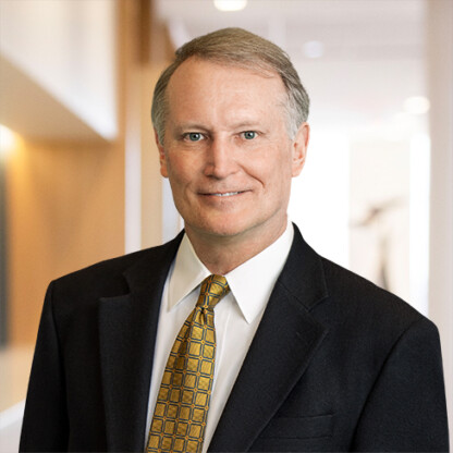 A middle-aged man in a dark suit and yellow patterned tie stands in a brightly lit hallway of a corporate law office, facing the camera and smiling slightly.